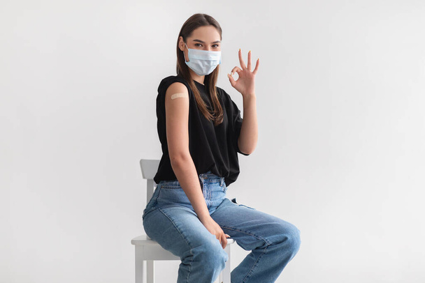 Giovane donna caucasica in maschera facciale dopo immunizzazione covid-19, mostrando spalla con cerotto, gesticolando bene - Foto, immagini