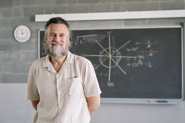 Portret van een vriendelijke seniorenleraar met baard in de klas met schoolbord met wiskundegraphics op de middelbare school. Onderwijs en opleiding Beroepsopleiding - Foto, afbeelding
