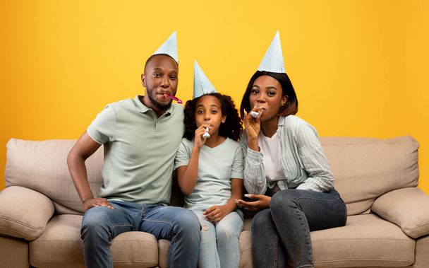 Χαρούμενοι Αφροαμερικανοί γονείς και κορίτσι γιορτάζουν τα γενέθλιά τους και φυσάνε στα κέρατα του πάρτι πάνω από κίτρινο φόντο - Φωτογραφία, εικόνα