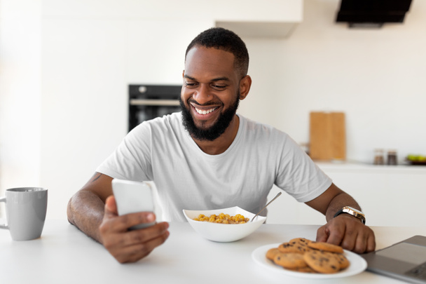 Hombre negro mensajes de texto en su teléfono inteligente desayunando en casa - Foto, imagen