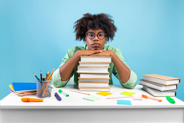 Lack of motivation. Bored african american teen guy sitting at table and leaning on stack of books, blue background - Fotó, kép