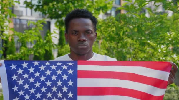 Крупным планом афро-американец держит американский флаг и смотрит в камеру летом - Кадры, видео
