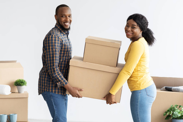 Home Relocation Concept. Joyful black spouses carrying cardboard boxes in their house - Photo, image
