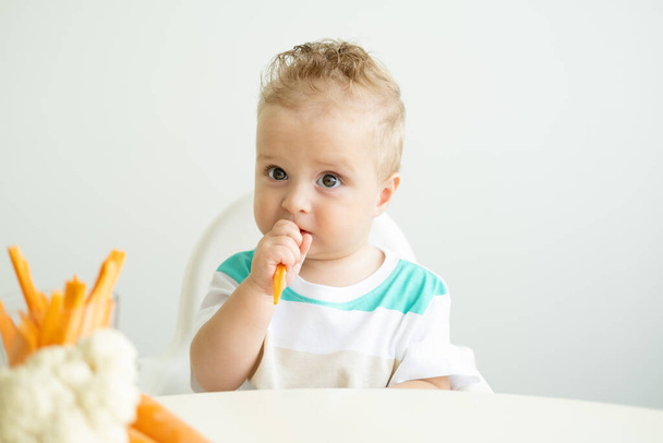 Bambino seduto in una sedia Childs mangiare fette di carota su sfondo bianco - Foto, immagini