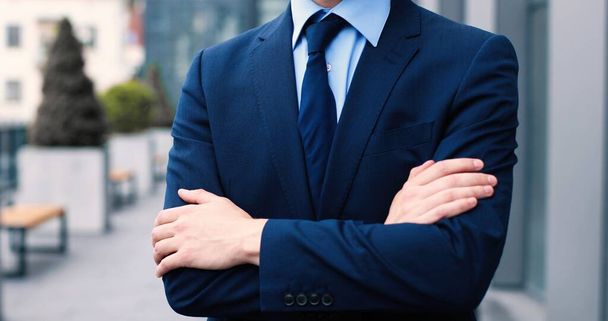 Confident and successful. Waist-up portrait of young elegant caucasian businessman is standing with crossed arms while looking at the camera with slight smile with his colleagues at the background - Photo, image