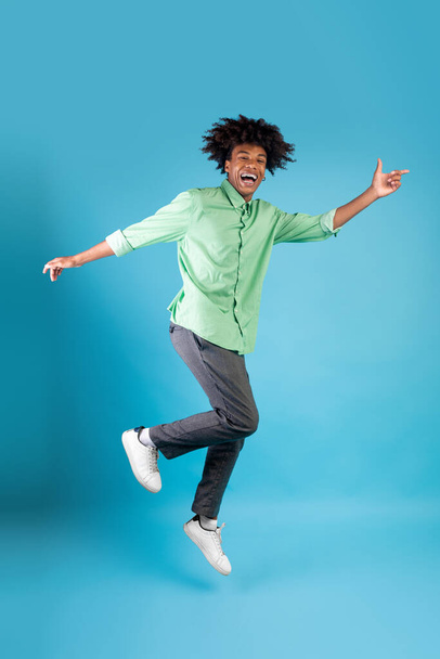 Joyful black guy jumping up in the air and showing funny figures, showing positive emotions over blue background, crop - Foto, afbeelding