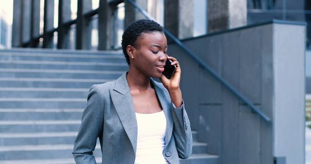 Attractive, good-wearing adult woman in suit standing nearby business center. She looking aside, smiling and holding telephone in hand while having pleasure conversation - Photo, image