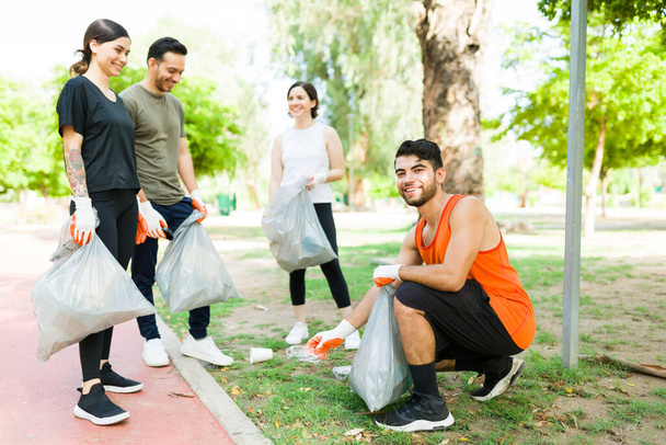 Schöner hispanischer Mann, der Müll im Park aufsammelt, bevor er gemeinsam mit einer Gruppe verantwortungsbewusster Freunde trainiert - Foto, Bild