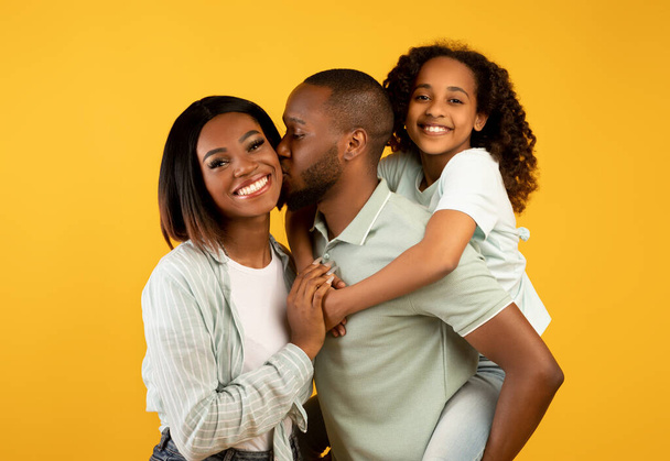 Glückliche afrikanisch-amerikanische dreiköpfige Familie mit entzückender Tochter, posiert und lächelt vor gelbem Hintergrund in die Kamera - Foto, Bild