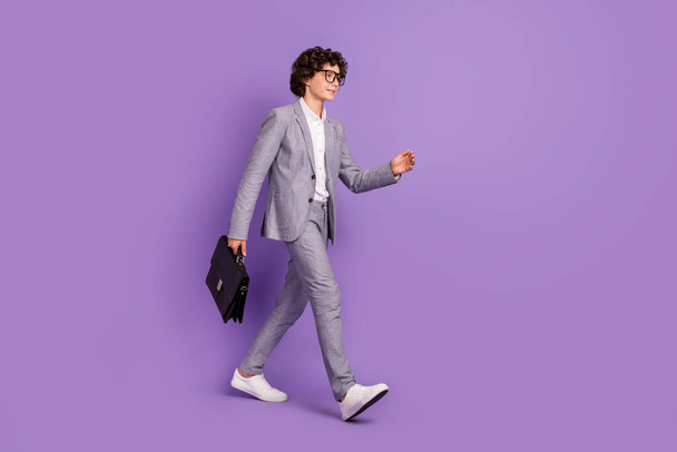 Fotografie pohledného sebevědomý žák chlap chůze tah nést pouzdro nosit šedý oblek obuv izolované fialové barvy pozadí - Fotografie, Obrázek