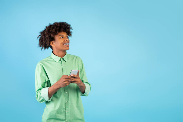 Mobile offer. Happy african american teen guy holding smartphone and looking aside at free space on blue background - Foto, afbeelding