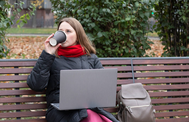 Mädchen arbeitet auf Parkbank am Computer und trinkt Kaffee - Foto, Bild