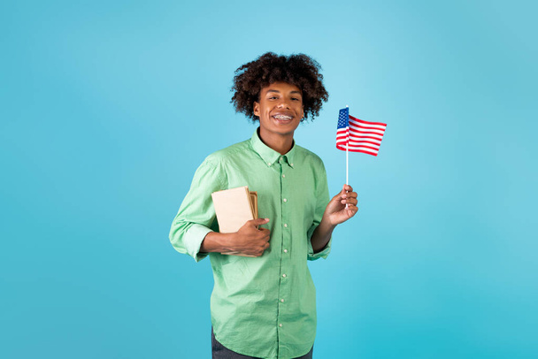 Student exchange and learning english. Zoomer happy black guy with books and small flag of USA, blue background - Foto, Imagem