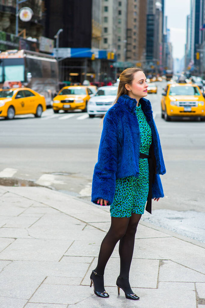 Gekleed in een blauwe nepbontjas, een jurk met patronen, zwarte legging en leren schoenen, staat een mooie vrouw in een drukke straat van een grote stad en kijkt om zich heen - Foto, afbeelding