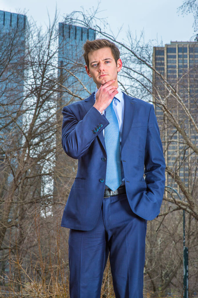 Dressing a blue suit and tie, one hand touching his chin , a young businessman is standing outside and thinking. The background is high business buildings - Photo, Image