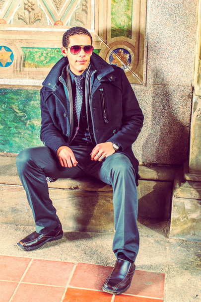 Jongeman, in de winter tegen de muur zittend, denkend, draagt wollen jas, stropdas, lange sjaal, zwarte broek, leren schoenen - Foto, afbeelding