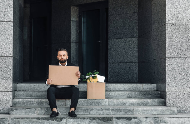 Werkloosheidsconcept. Depressieve volwassen zakenman zittend met leeg kartonnen bord en doos met persoonlijke bezittingen - Foto, afbeelding