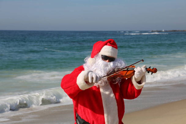 Noel zamanı. Noel Baba. Keman. Kemancı. Noel Baba sahildeyken keman çalar. Noel Baba sahilde tatildeyken kemanını çalar. Noel Baba müzik çalar. Noel Baba plajda müzik konserinde. Noel müziği. Ho Ho Ho. Mutlu Noeller.. - Fotoğraf, Görsel