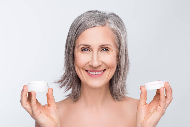 Photo of positive cheerful old woman hold hands cream smile lifting hydration isolated on grey color background - Foto, Imagem