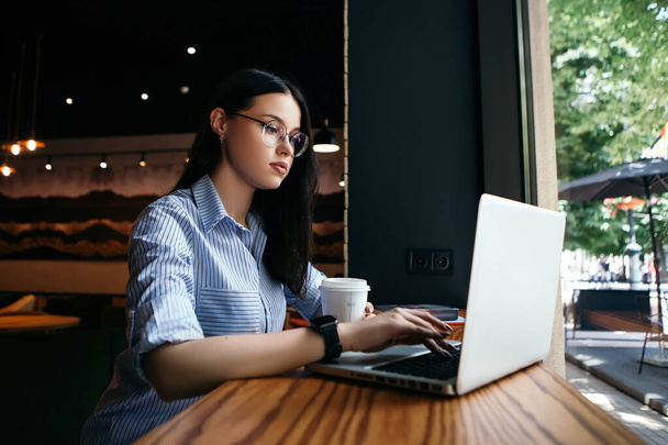 ビジネス女性のフリーランサーカフェドリンクホットドリンクコーヒーブレイクは、ランチ作業ノートパソコンの入力メッセージテキストスマートウォッチ手はスタイリッシュなメガネコピースペースを着用して座っている。国際コーヒーの日!  - 写真・画像