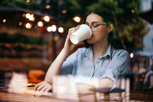 Lövés az ablakon keresztül üzlet nő szabadúszó ül kávézó ital forró ital kávészünet ebéd előtt munkanap okos óra kéz viselni elegáns szemüveg másolás. Nemzetközi Kávénap!  - Fotó, kép