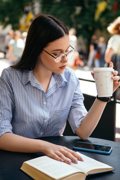 Business woman sitting outside terrace porch cafe drink hot beverage coffee break have lunch reading book smart watch hand wear stylish glasses copyspace. Rush hour, International Coffee Day  - Фото, изображение