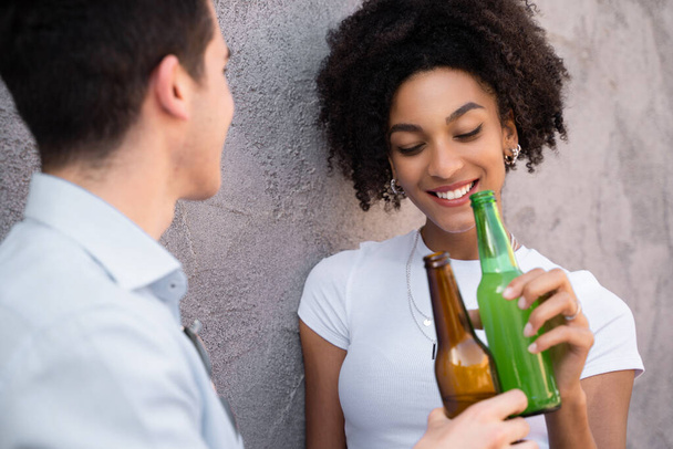 Багаторасова пара молодих закоханих розмовляє між собою, тримаючи і випиваючи пляшки пива. Сором'язлива афроамериканка з закритими очима.. - Фото, зображення
