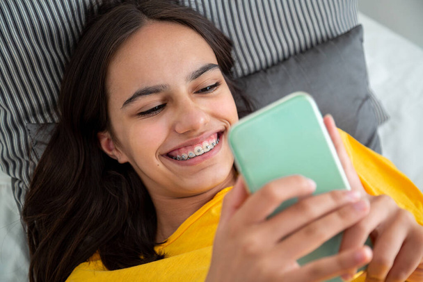 Happy teen girl with braces using her smartphone lying in bed smiling - Foto, Imagen