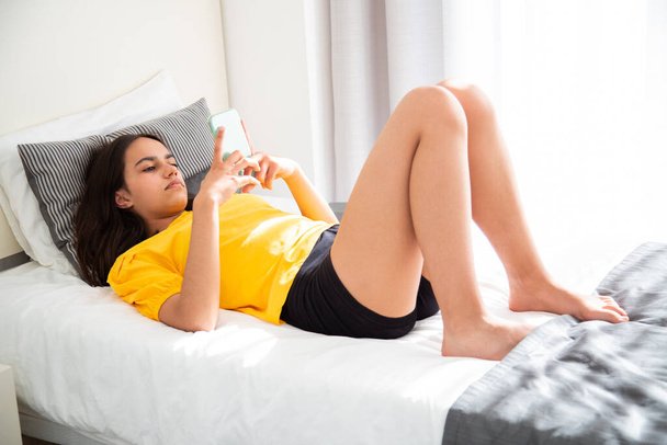 Teen girl wearing a yellow t-shirt focused on her smartphone lying in bed - Фото, зображення