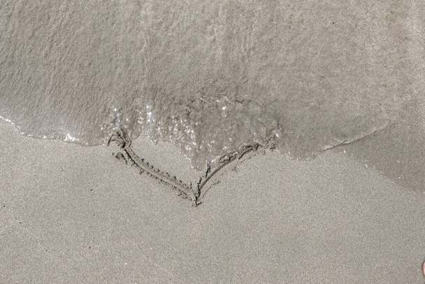 Sea waves wipe hand drawing heart shape in the sand on the beach. Summer love concept. Close up, copy space for design - 写真・画像