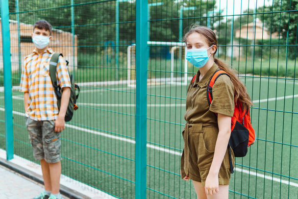 dos adolescentes niño y niña en el camino a la escuela, que utilizan mascarillas protectoras, de pie junto al patio de recreo y mostrar la distancia social para proteger contra la infección coronavirus - Foto, imagen