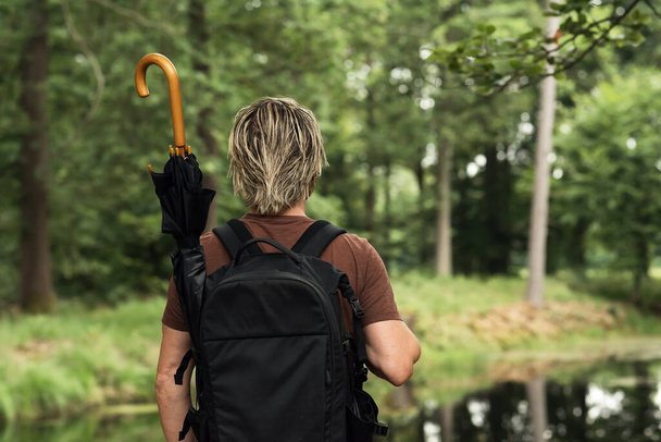 Hombre rubio en camiseta marrón con una mochila con un paraguas en ella se encuentra en un bosque cerca de un estanque. Vista trasera. - Foto, imagen