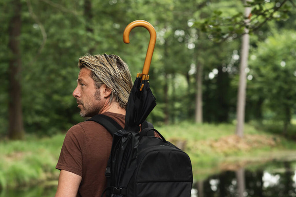 Blonde man with stubble beard in brown t-shirt with a backpack with an umbrella in it looking over his shoulder stands in a forest near a pond. Rear view. - Fotó, kép