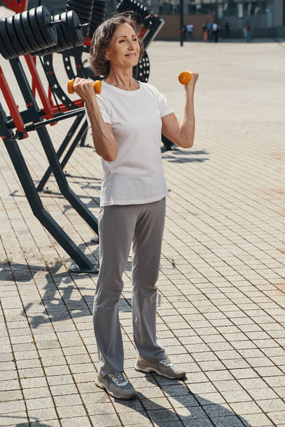 Cheerful aged woman exercising arms with weights - Photo, Image