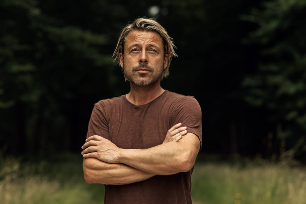 Blonde man with his arms folded and a stubble beard in a brown t-shirt in a forest in summer. Front view. - Photo, Image