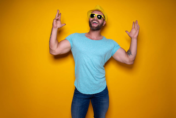Happy male in blue shirt, sunglasses and yellow hat celebrating success standing on yellow background.  - Foto, immagini
