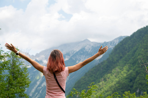 Kilátás hátulról egy fiatal nő áll odakint zöld természetben nézi hegyek széttárt karokkal.. - Fotó, kép