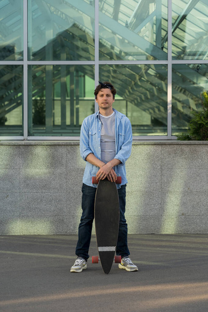 Stylish man holding longboard over city building. Hipster guy skateboarder with skateboard outdoors - Foto, imagen