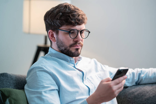 Симпатичный парень сидит дома на диване и держит телефон в руках, переписывается с друзьями в чате - Фото, изображение