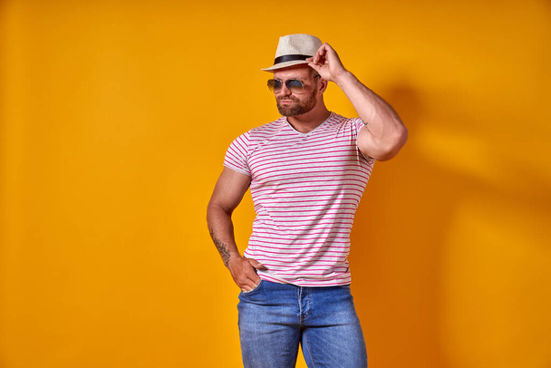 Muscular bearded male in shirt, jeans and stylish hat, sunglasses standing in studio over yellow background - 写真・画像