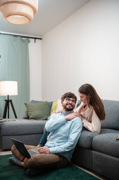 Cuidar da esposa dá ao marido uma massagem no ombro enquanto ele trabalha remotamente no laptop easys em casa, sentado no chão - Foto, Imagem