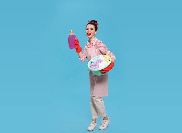 Housewife holding bottle of cleaning product and basin with clothes on light blue background - Φωτογραφία, εικόνα