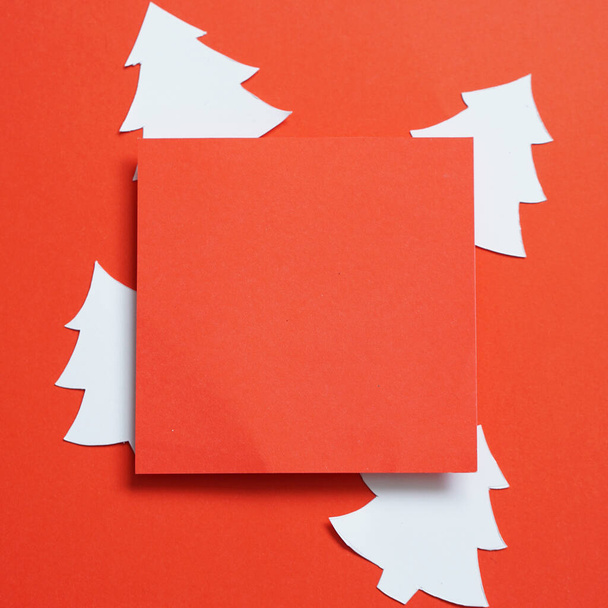 Рождественский фон с пустой красной квадратной поздравительной открыткой из бумаги и белой бумаги, вырезанной в форме рождественской елки на красном фоне. - Фото, изображение