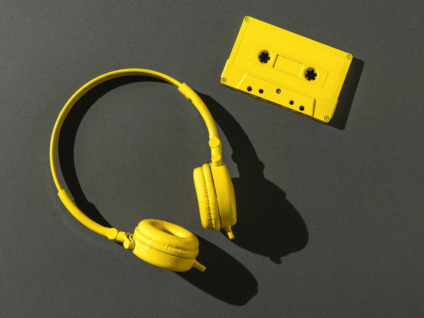 Koptelefoon en een gele cassette met een magneetband op een zwarte achtergrond in fel licht. Kleurtrend. Vintage apparatuur voor het luisteren naar muziek. Vlakke plaat. - Foto, afbeelding