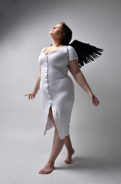 Full length πορτρέτο της νεαρής συν μέγεθος γυναίκα με κοντά καστανά μαλλιά, φορώντας ένα σφιχτό λευκό φόρεμα και την έλλειψη φτερό φτερά αγγέλου, στέκεται ποζάρουν με σκιερό φωτισμό backlight στο φόντο στούντιο. - Φωτογραφία, εικόνα