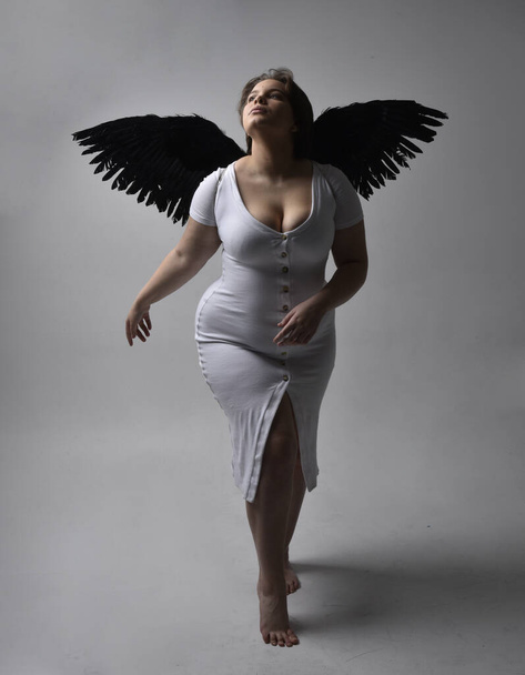 Full length portrait of young plus sized woman with short brunette hair,  wearing a tight white  dress and lack feather angel wings, standing pose with shadowy backlit lighting on studio background. - Fotoğraf, Görsel