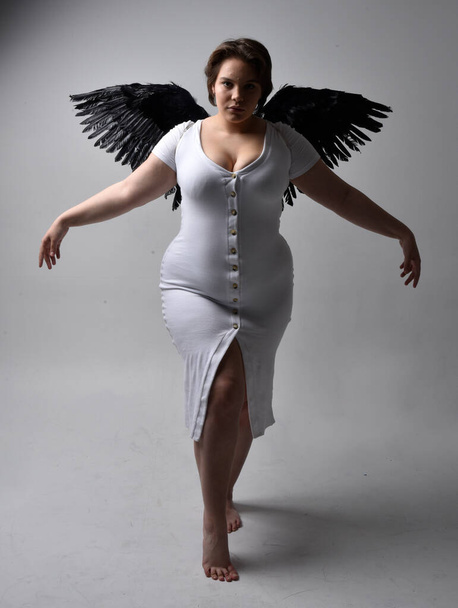 Retrato de larga duración de una joven mujer de tamaño grande con cabello corto y morena, con un vestido blanco ajustado y sin alas de ángel de plumas, posando de pie con iluminación retroiluminada sombría en el fondo del estudio. - Foto, imagen