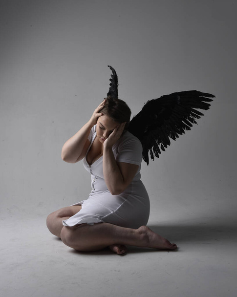 Full length portrait of young plus sized woman with short brunette hair,  wearing a tight white  dress and lack feather angel wings, kneeling pose with shadowy backlit lighting on studio background. - Photo, Image