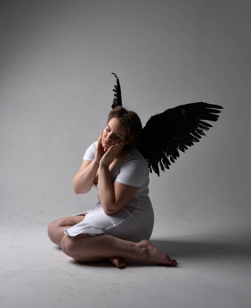 Full length πορτρέτο της νεαρής συν μέγεθος γυναίκα με κοντά καστανά μαλλιά, φορώντας ένα σφιχτό λευκό φόρεμα και την έλλειψη φτερό φτερά αγγέλου, γονατιστή στάση με σκιερό φωτισμό backlight στο φόντο στούντιο. - Φωτογραφία, εικόνα