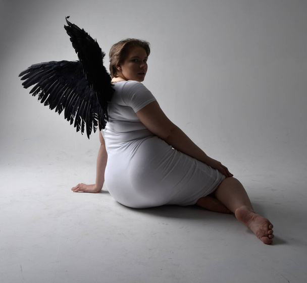 Full length portrait of young plus sized woman with short brunette hair,  wearing a tight white  dress and lack feather angel wings, kneeling pose with shadowy backlit lighting on studio background. - 写真・画像
